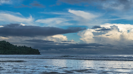Morning at the ocean. The blue clouds in the azure sky are highlighted. The waves are rolling towards the shore. A hill in the distance. Malaysia. Borneo. Kota Kinabalu.