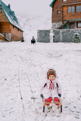 Little smiling girl slides down a snowy hill on a sled against the backdrop of her mom sitting near...