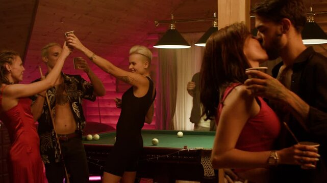 Young partygoers gather in a wooden room as a couple kisses passionately while holding cups, and friends clink paper cups behind them; a man dances by curtains, setting a festive, relaxed mood.
