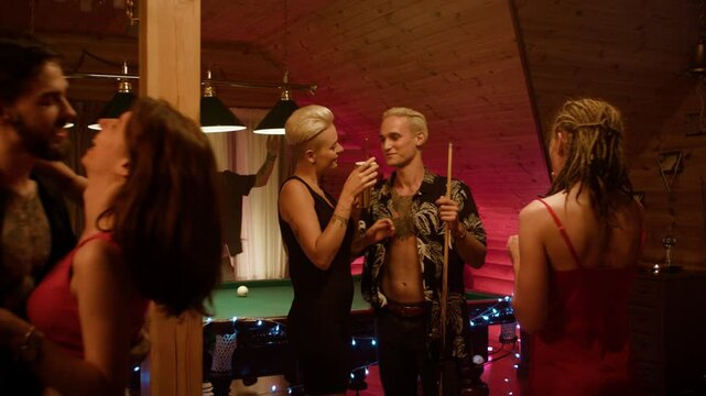 Wooden party: woman in a black dress with short light hair talks with a floral man while a red-top, white-shorts couple kisses upfront and a man in dark shirt dances by curtains amid scattered balls.
