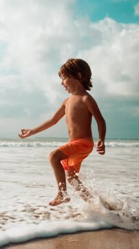 Vertical shot. Slow motion of barefoot little dreamer boy run and jump with arms up on beach. Concept of freedom, happy moment in childhood, kid have fun. Summer family trip on weekends to the sea