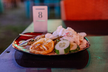 Indonesian food. Fried noodles with omelette and sliced cucumber on a dining table in a restaurant at night.