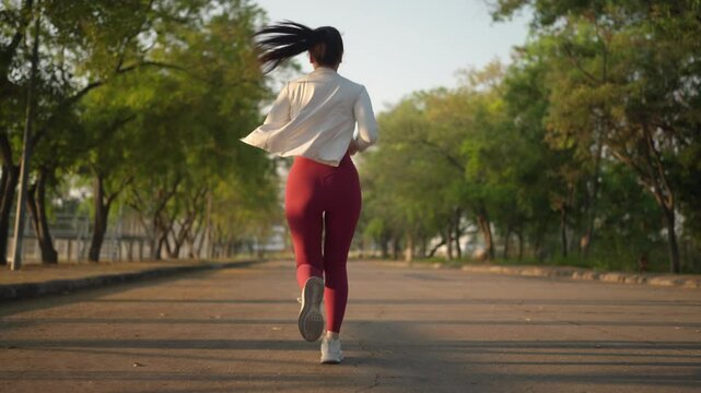 A woman is running on a road with trees in the background. She is wearing red pants and a white shirt