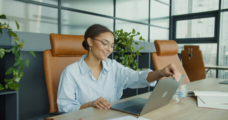 smiling young woman in glasses sitting at a modern office desk, closing her laptop while enjoying a...