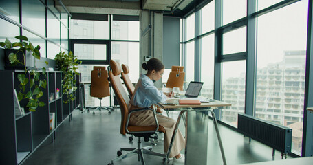focused young professional woman working on a laptop in a modern office, analyzing architectural plans with notes on a notepad, surrounded by natural light and urban views