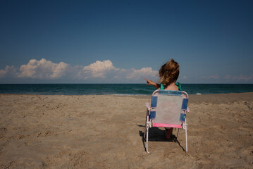 Toddler girl sitting in beach chair pointing at ocean waves