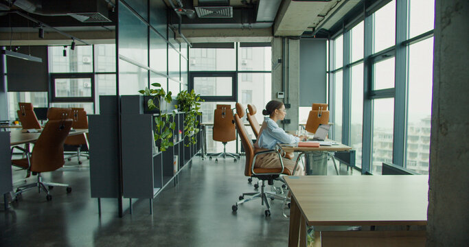 A focused professional woman working on a laptop at a modern office desk, taking notes in a notebook, surrounded by stylish furniture, plants, and natural light from large windows