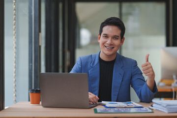 Fototapeta premium Happy young manager giving a thumbs up while working at a desk with a laptop and business charts, celebrating the success of a startup project and enjoying the journey