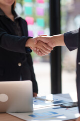 Businesswomen shaking hands over a desk with a laptop and documents with charts and graphs,...