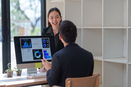 Two asian businesspeople are discussing business performance using charts and data displayed on a computer screen, working together in the office