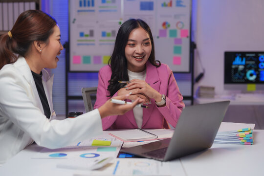 Two smiling asian businesswomen are discussing work, using a laptop, in a modern office meeting room with graphs and charts displayed on a whiteboard and computer monitor