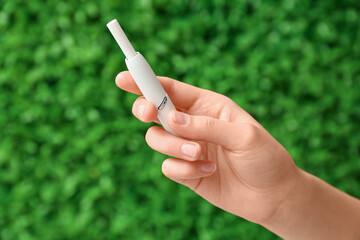 Female hand holding modern electronic cigar with stick against green nature background
