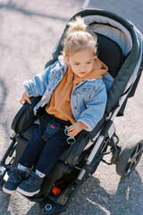 Little girl with sunglasses in her hand sits in a stroller and looks away