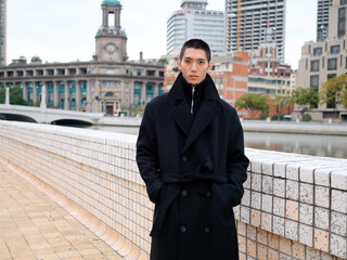 Portrait of handsome Chinese young man wearing black overcoat posing in the street, young guy with black short hair with Shanghai bund background. Male fashion, cool Asian young man lifestyle.