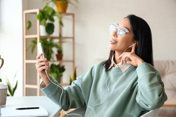 Young Asian woman in earphones using mobile phone at home office