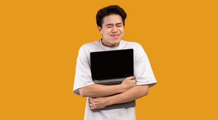 Technology Lover Concept. Happy excited asian man hugging laptop with black blank empty screen, holding it tight near chest. Geek posing isolated over orange studio background wall, copy space