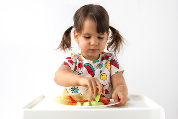 Little girl with pigtails sitting in high chair, carefully picking up  piece of cucumber from plate of fresh vegetables. Healthy eating, childhood nutrition,  independent eating