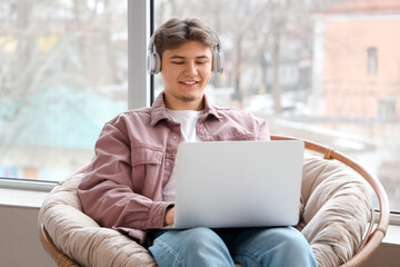 Male student with headphones and laptop studying online in armchair at home