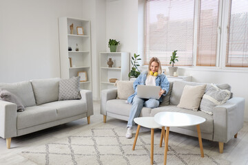 Young woman in headphones sitting on sofa and using laptop in light living room