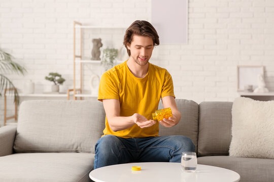 Handsome young happy man with jar of fish oil capsules and water sitting on sofa at home