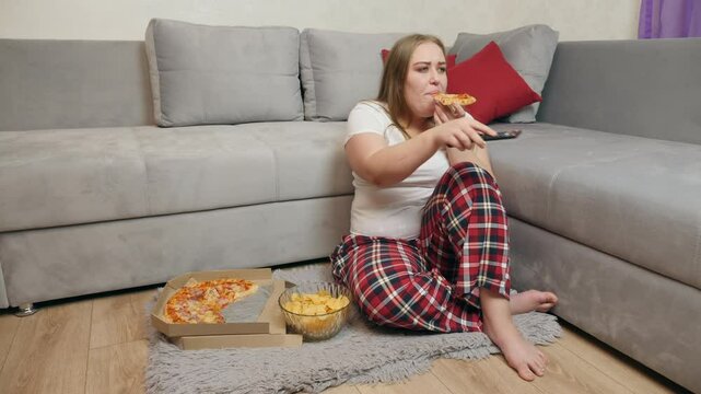 A woman in pajamas sits on the floor near the couch, holding a slice of pizza and a TV remote. On the rug in front of her are a pizza box and a bowl of chips