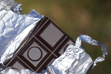 Closeup broken Dutch dark chocolate bar in silver foil. Blurred garden as background.
