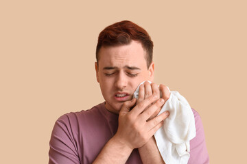 Young man with cold compress suffering from toothache on beige background