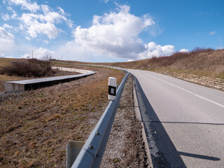 A curving rural road with a guardrail stretches uphill under a bright blue sky. A drainage channel...