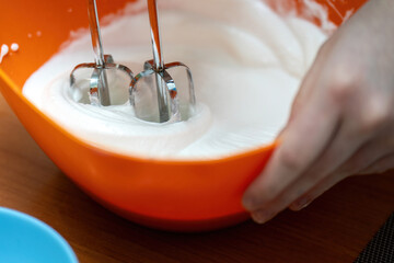 metal mixer whisks beating a translucent egg white into a stiff white foam, set against a vibrant orange bowl, showcasing the process of baking and food preparation