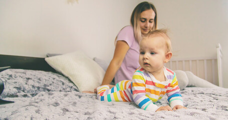 A young mother looks at her phone while her baby crawls on the bed next to her.
