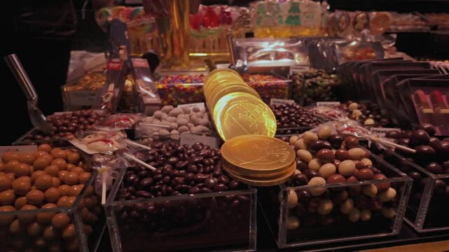 Close-up of a chocolate and candy stall at Mercat de la Boqueria in Barcelona, featuring chocolate-coated nuts, sweets, and golden foil-wrapped chocolate coins under warm lighting.