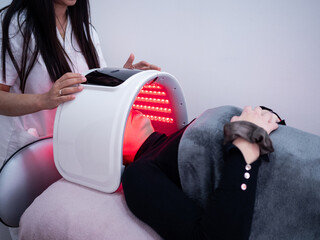 A woman enjoys a soothing LED light mask therapy session at a wellness center. A therapist adjusts...