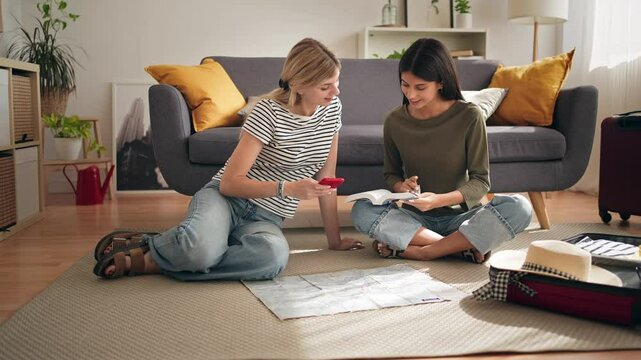 Female friends planning a trip together, looking at a guidebook and at a mobile phone at home