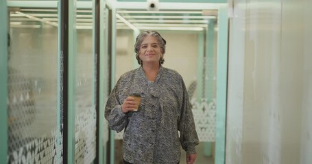 Indian gen x senior old aged boss woman lady ceo hold paper cup walk indoor office space smiling elder older active female staff worker drink coffee spend day time break hall way job work place