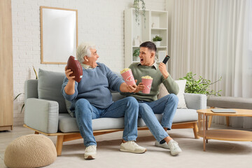Young man and his father with popcorn watching rugby game on TV at home