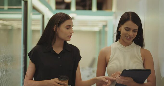 Indian young adult gen z woman lady use tablet walk office space talk work task two expert girl staff hold paper cup drink coffee speak casual enjoy break spend day time at job place have fun joy