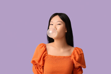 Pretty young Asian woman blowing bubble gum on lilac background