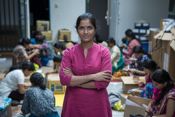 an indian woman confidently standing with arms crossed, surrounded by a group of other women who appear to be working on a project.