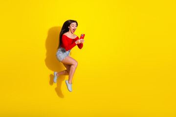 Energetic young woman jumping joyfully while using her red smartphone against a vibrant yellow background