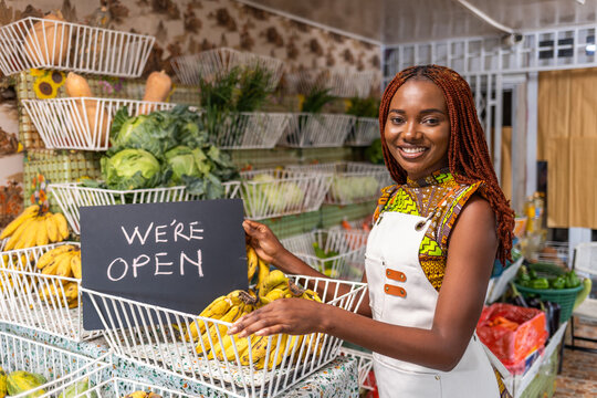 Successful Black female African entrepreneur holding "we are open" sign in front of her grocery shop where she sells healthy fresh fruits and vegetables, in a local market.