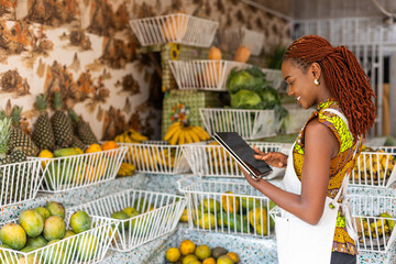 Successful African female business owner of fresh produce store and grocery shop uses a smart tablet to video call customers and take orders remotely.