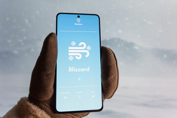 Hand wearing a glove holding a smartphone displaying a weather app with a blizzard warning. Snowy landscape and strong wind in the background. Extreme winter weather and forecast technology concept