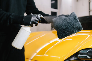 Car cleaner spraying liquid for waxing on microfiber cloth at the service. Detailing process with...
