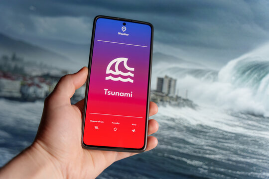 Hand holding a smartphone displaying a weather app with a tsunami warning. Giant ocean waves approaching a coastal city in the background. Natural disaster and emergency alert concept