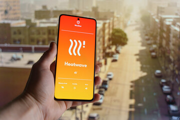 Hand holding a smartphone displaying a weather app with a heatwave warning. Blurred cityscape under extreme heat in the background. High temperature and climate change concept