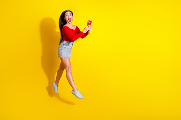 Young woman in a vibrant outfit joyfully jumping while holding a smartphone against a bright yellow background