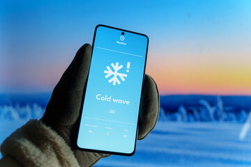 Hand wearing a glove holding a smartphone displaying a weather app with a cold wave warning. Extreme cold and weather forecast concept