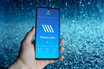 Hand holding a smartphone displaying a weather app with a heavy rain forecast. Raindrops falling in...