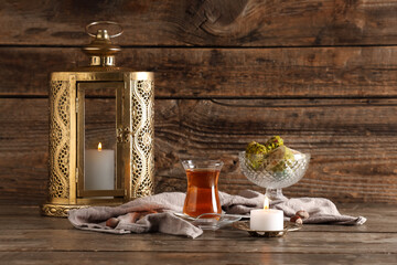 Glass of tasty Turkish tea with fanoos, burning candle and sweets on wooden background. Ramadan...