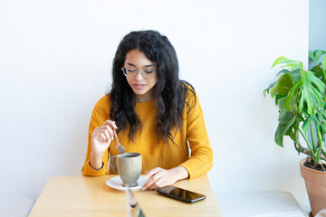 Latin woman stirring coffee in cafe with smartphone nearby